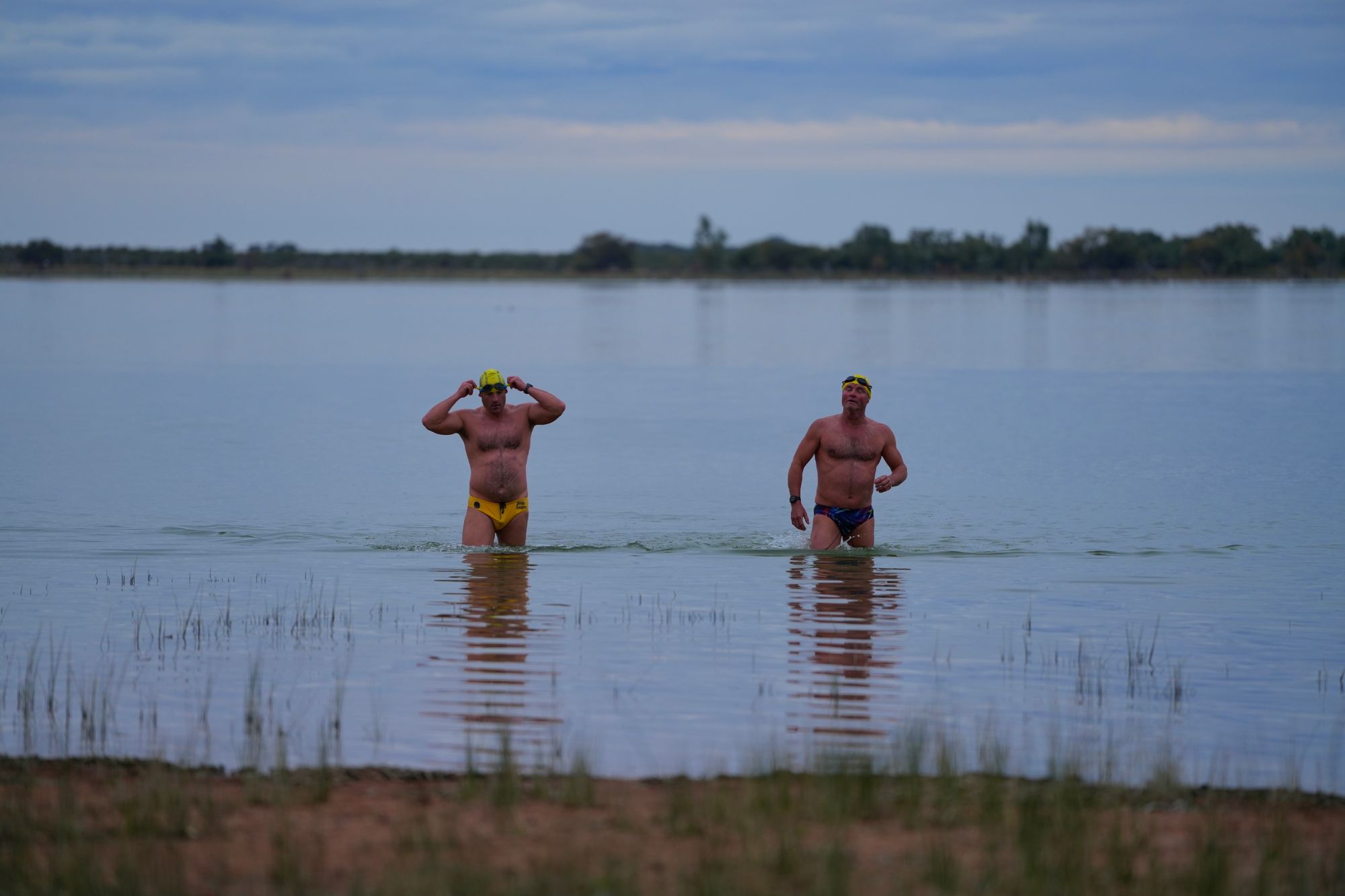 Two men in swimming outfits exit a lake.