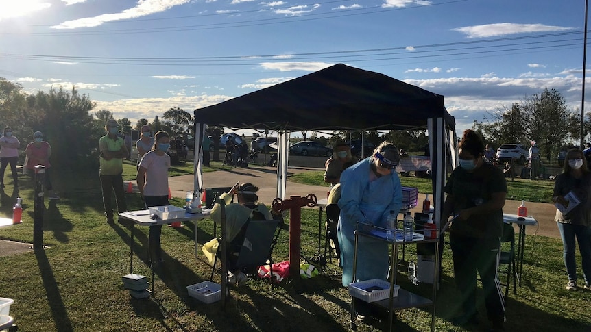 People wearing blue suits and plastic face shields standing under a gazebo. People wearing face masks lined up in front.