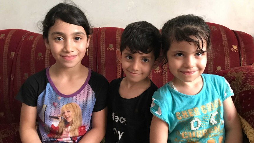 Three children smile as they sit on a couch in their home.