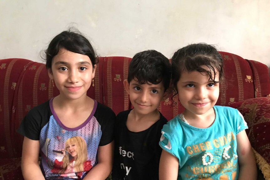 Three children smile as they sit on a couch in their home.