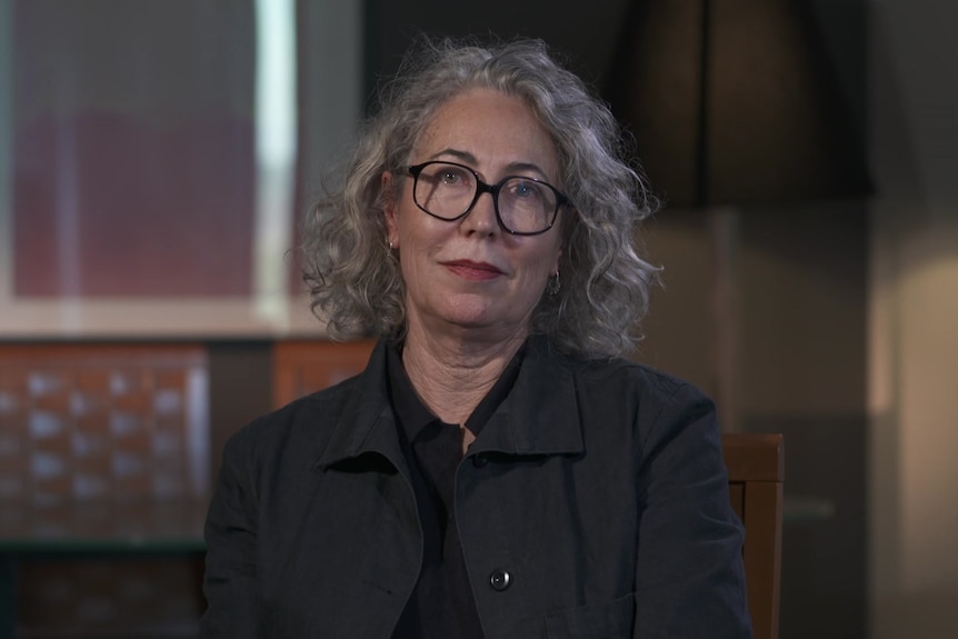 A woman wearing glasses looks into camera with a serious expression.