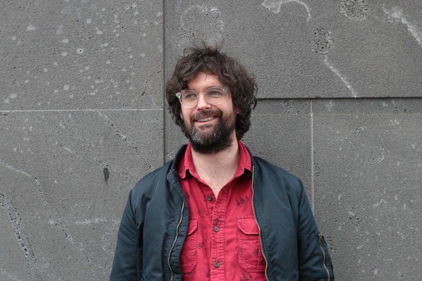 A white man with a unkempt beard and wild hair with a cement wall in the background