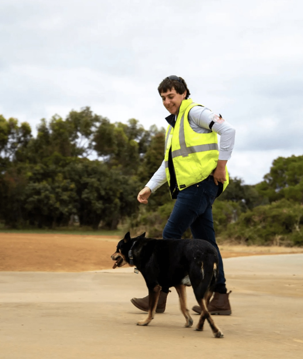 A young man with a high vis vest walks on an airfrield next to his dog.