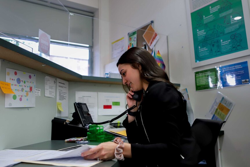 Woman with black hair and black jacket holds telephone looking at papers sitting at desk inside office