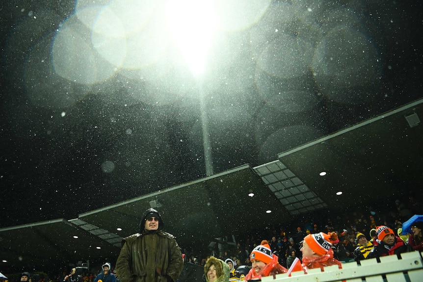 Fans are rugged up as the floodlights beam down and snow falls.