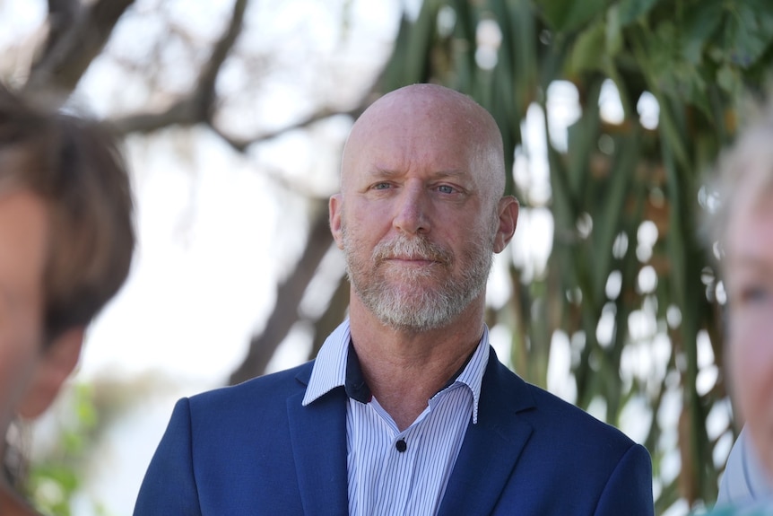 Bald man with short grey beard wearing blue striped shirt and navy blazer staring ahead