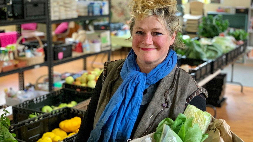 a woman wearing a blue scarf holds a parcel of fruit, vegetables and dry goods, with shelves of food behind her.