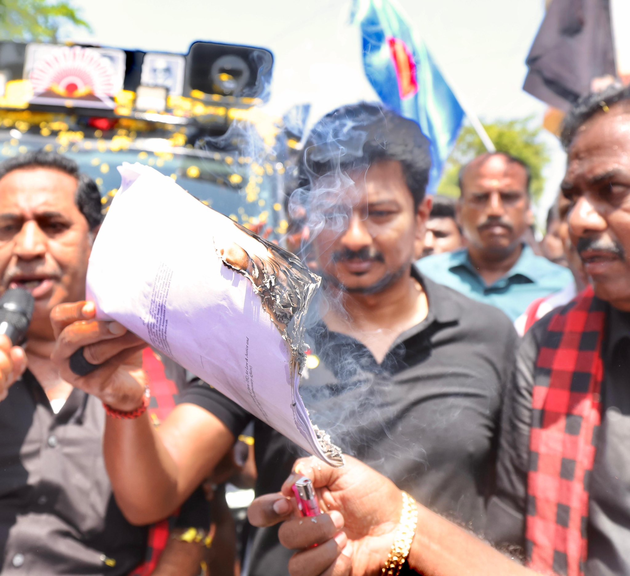 A crowd of protesters gather around a man in a black t-shirt burning a piece of paper