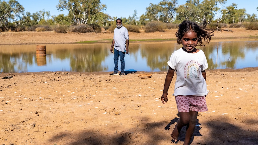An Indigenous man standing by a river, watching a young girl walk away.