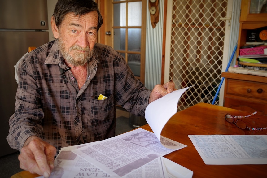 A man sitting at a kitchen table looking at the camera, holding stack of papers