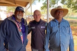 three men smiling, standing in remote community with red dirt