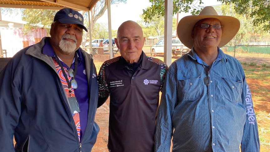 three men smiling, standing in remote community with red dirt