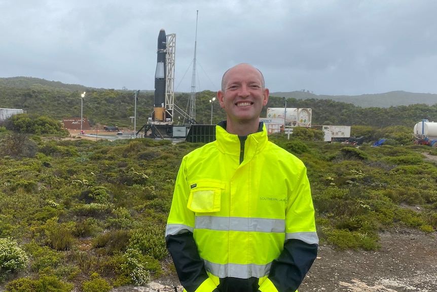Smiling man in high-vis jacket standing in front of rocket site with tanks, green bushes