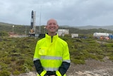 Smiling man in high-vis jacket standing in front of rocket site with tanks, green bushes