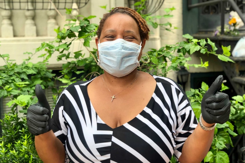 A young black woman in black and white summer dress and face mask gives thumbs-up sign on sunny day.