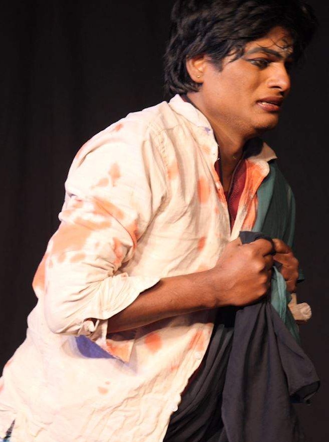 An Indian man in what appears to be a blood-spattered shirt, performs onstage, looking fearful as he clutches his belongings.