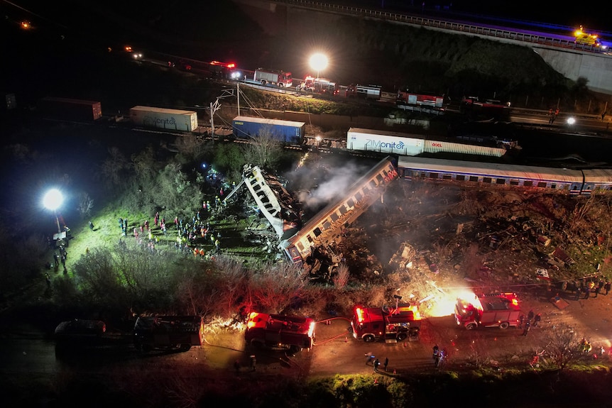 An aerial view showing a train crash site illuminated by spotlights and emergency sirens. Several carriages are off the tracks.
