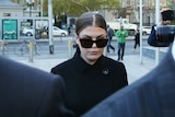 Wearing dark glasses and a dark jacket, Belle Gibson can be seen arriving at court behind the shoulders of two men in suits.
