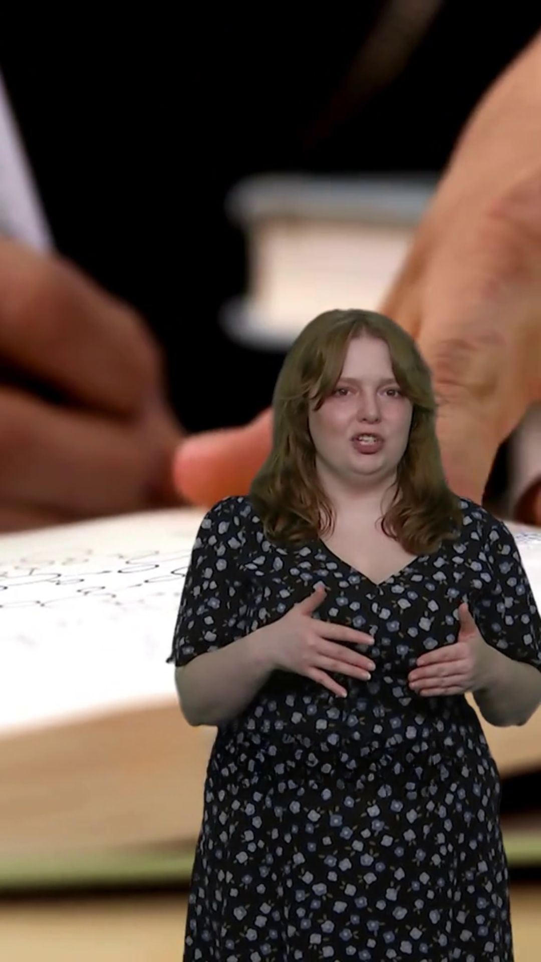 A red-headed woman speaks to camera. She wears a black and blue floral dress. 