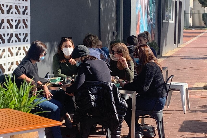 A group of women sit at a cafe table wearing masks outside.