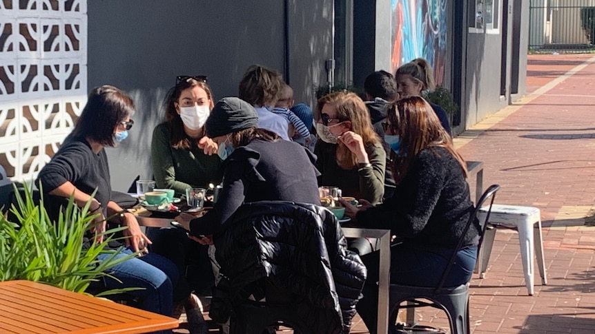 A group of women sit at a cafe table wearing masks outside.