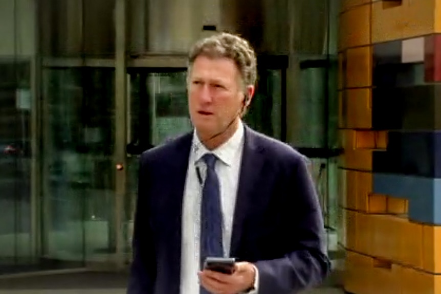 Johnathan Kenny, wearing a suit and tie with headphones in, leaves the orange Federal Court building.