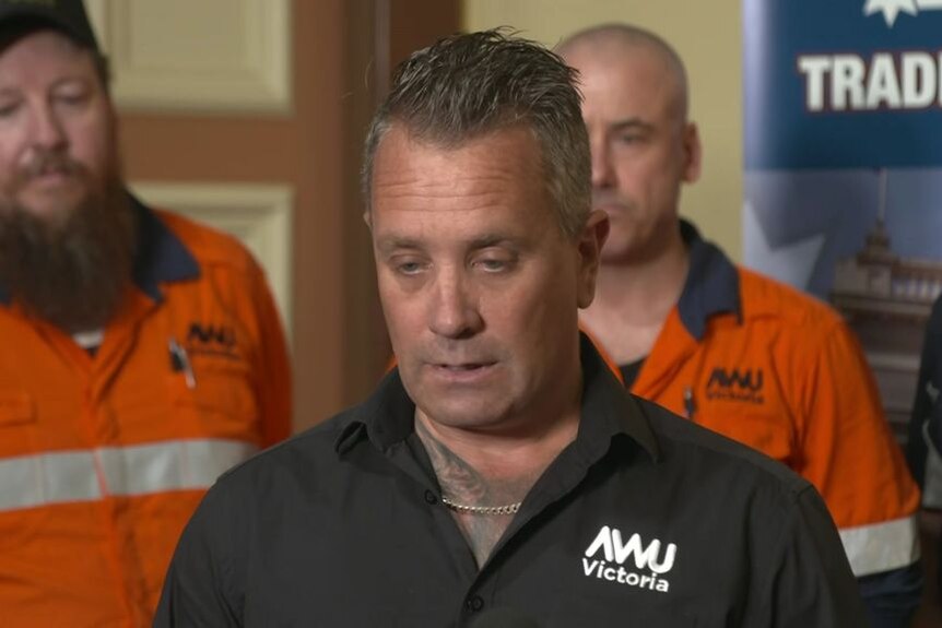 A man with an AWU Victoria shirt gives a media conference. Two men in orange high-vis stand behind him.