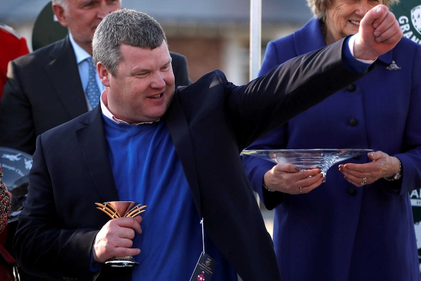 A man in a suit jacket and blue jumper punches the air after being presented with a trophy.