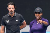 A man wearing black stands behind a female tennis player who is pointing towards the camera