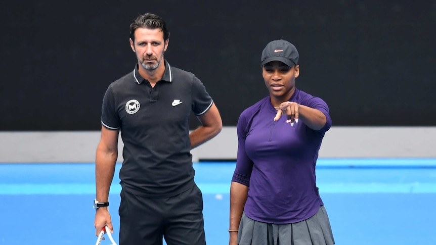 A man wearing black stands behind a female tennis player who is pointing towards the camera