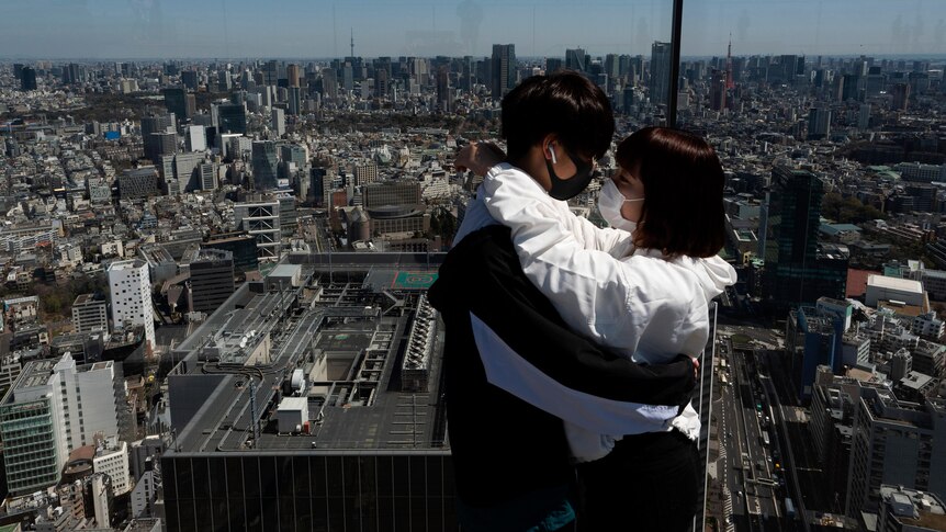 Two people in facemasks hug each other on an observation deck with a city horizon extending into the background