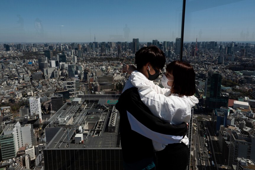 Two people in facemasks hug each other on an observation deck with a city horizon extending into the background