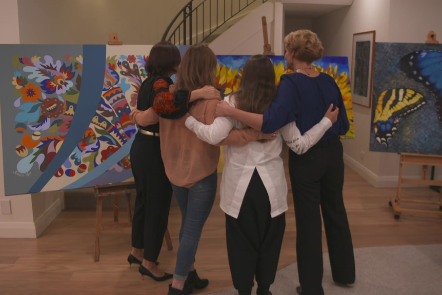 Four women with their arms around each other, standing in front of artworks.