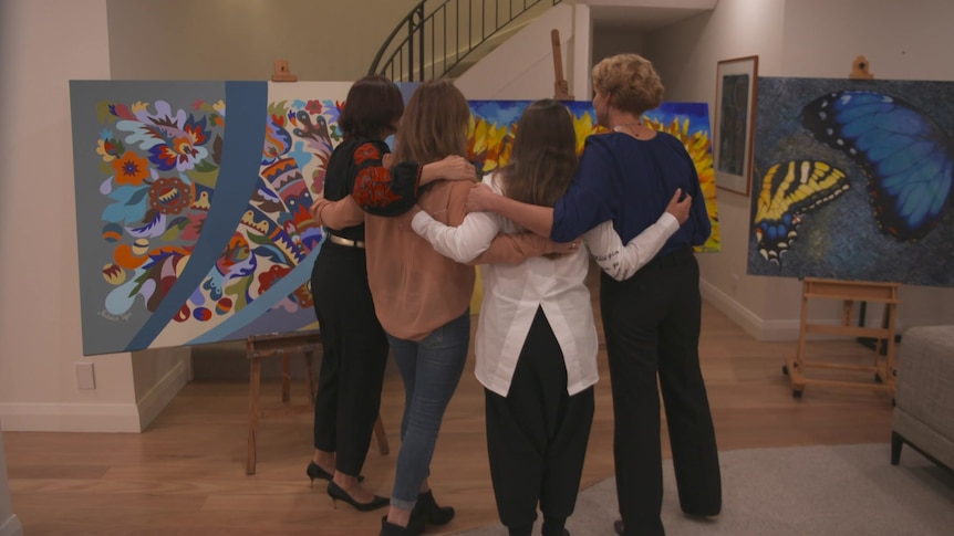 Four women with their arms around each other, standing in front of artworks.