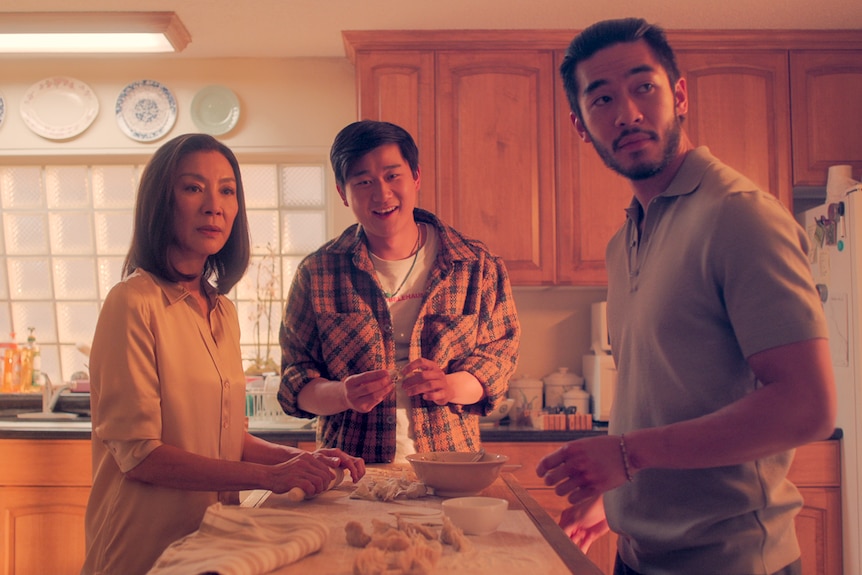 The family stand around a kitchen table in a dated room and look behind the camera with serious, happy and concerned faces