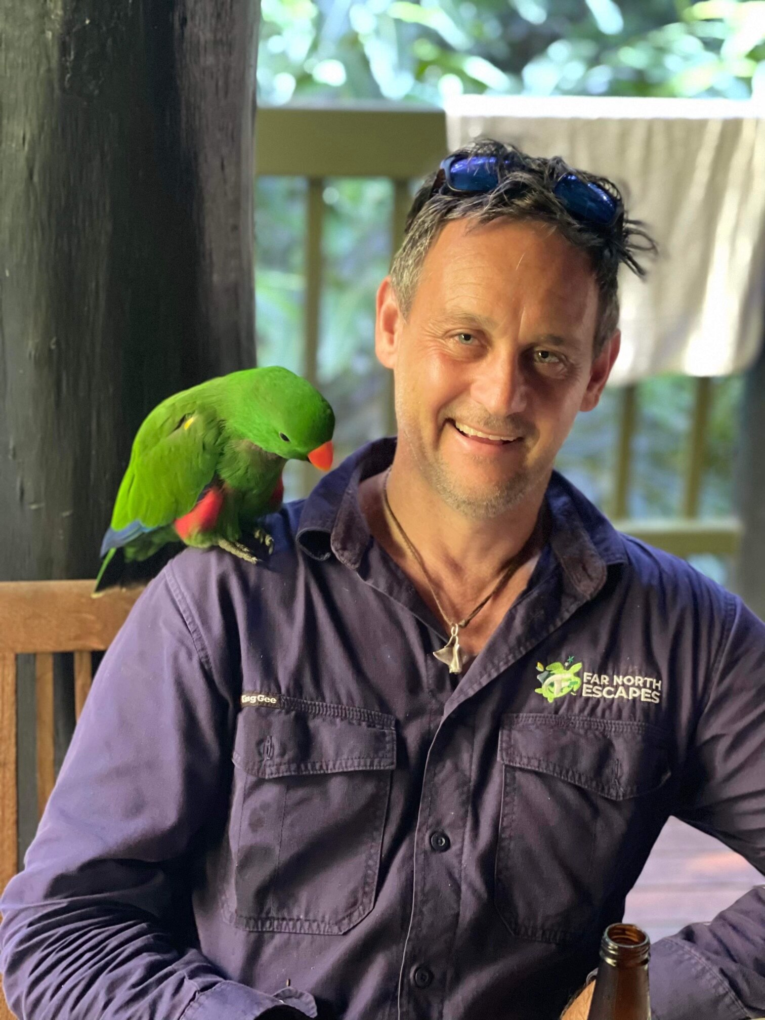 Mark Cromwell wearing a "Far North Escapes" business shirt and smiling, with a green tropical parrot sitting on his shoulder.