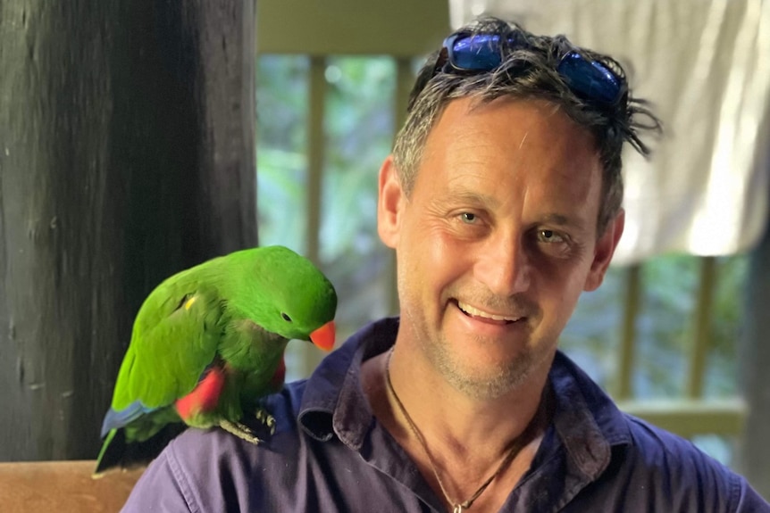 Mark Cromwell wearing a "Far North Escapes" business shirt and smiling, with a green tropical parrot sitting on his shoulder.