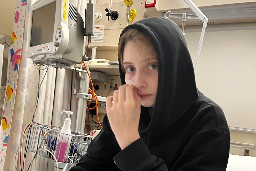 A young teenager wearing a hoodie, sitting on a hospital bed. He is looking at the camera with a concerned expression.