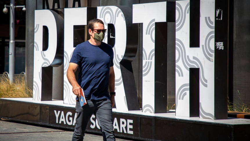 A man wearing a mask walks past a "Perth" sign