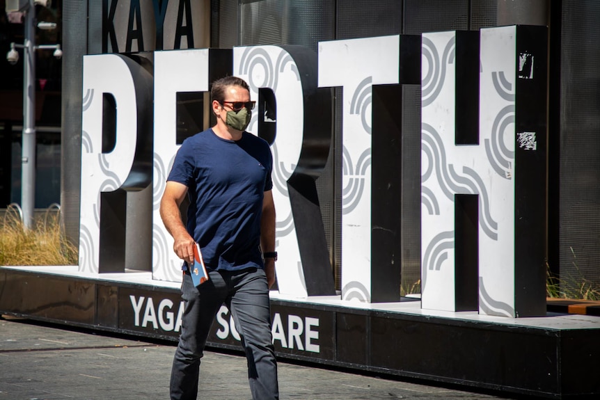 A man wearing a mask walks past a "Perth" sign