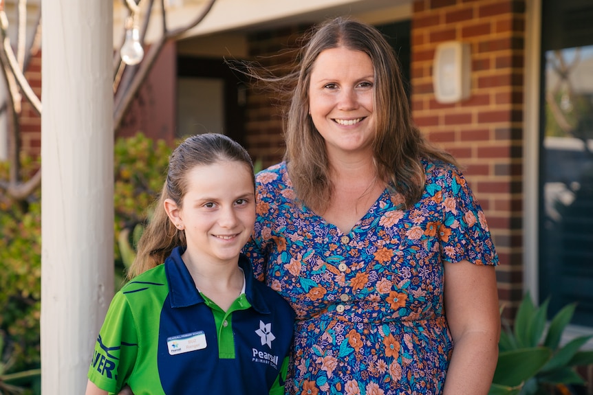 A young girl in her school uniform stands next to her mother outdoors, with both of them smiling for the camera.