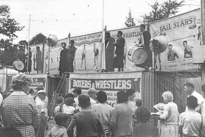 A crowd standing at a platform with advertising a boxing and wrestling tent.