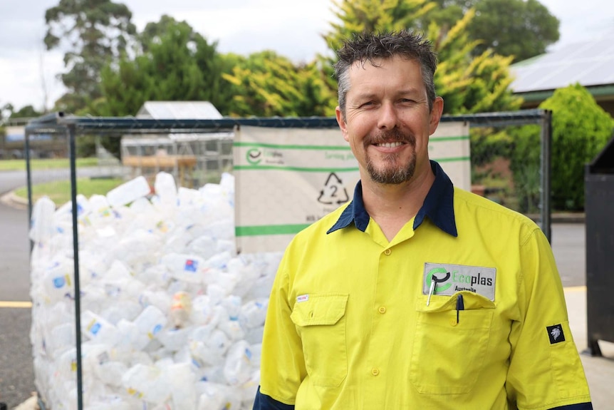 A man wearing a high vis shirt reading "Ecoplas" smiles in front of a big container of empty plastic milk bottles.