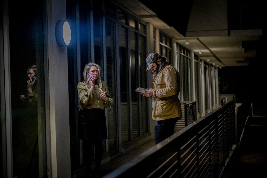 On a dark veranda, under a bright porch light, a woman talks into a mobile phone as a man writes in his notepad