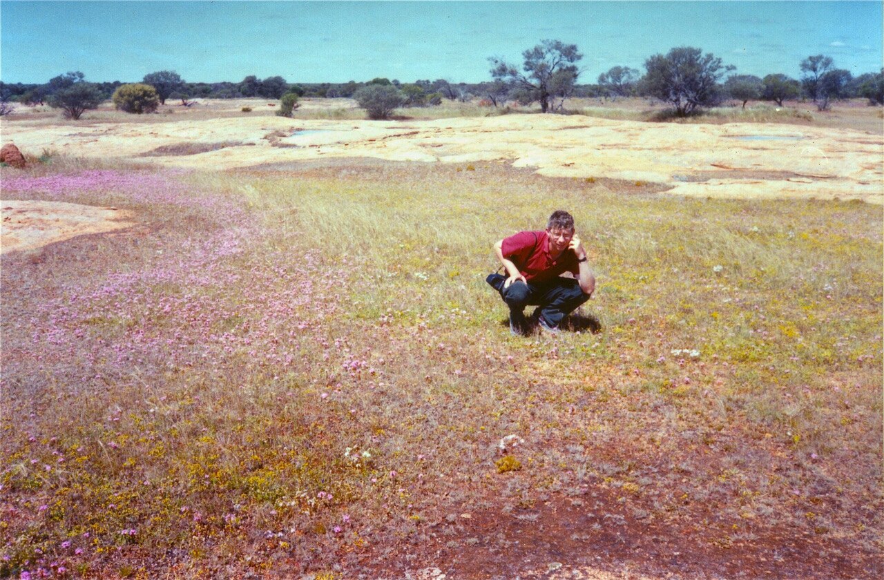 Jerry Coleby-Williams squats in a dry field with small purple flowers