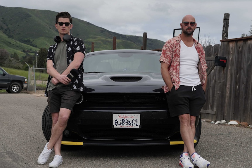 Two men leaning against the bonnet of a black muscle car.