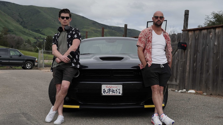 Two men leaning against the bonnet of a black muscle car.