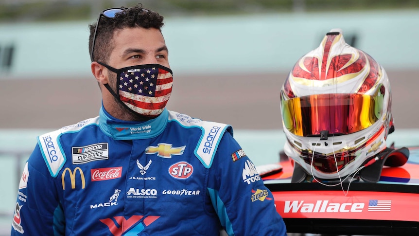A NASCAR driver stands in front of his car, while wearing an American flag face mask.