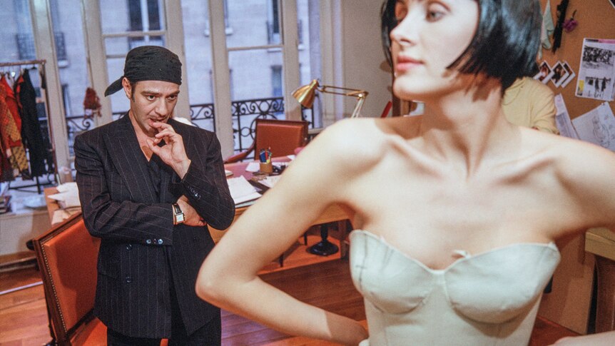 The designer John Galliano in his early 40s in black suit and hat, looking upon a model being fitted, in 2005