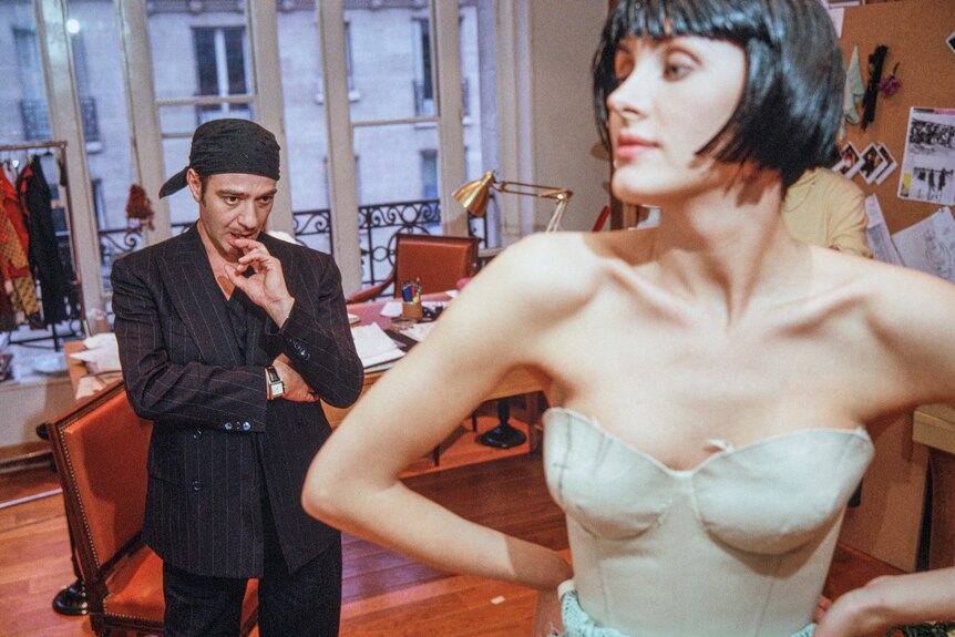 The designer John Galliano in his early 40s in black suit and hat, looking upon a model being fitted, in 2005